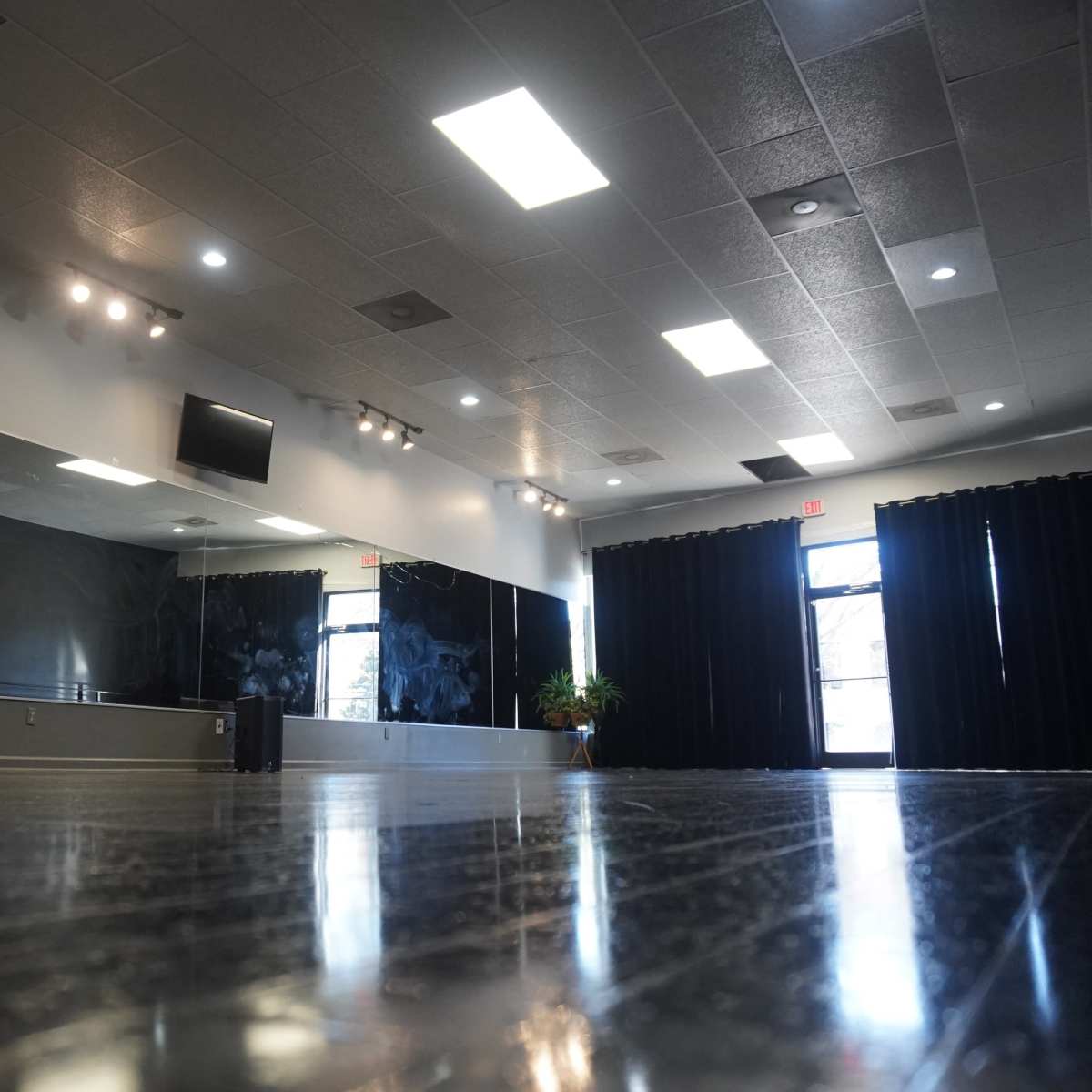 An empty dance studio with a mirrored wall, black curtains, and a wooden floor reflecting overhead lights.