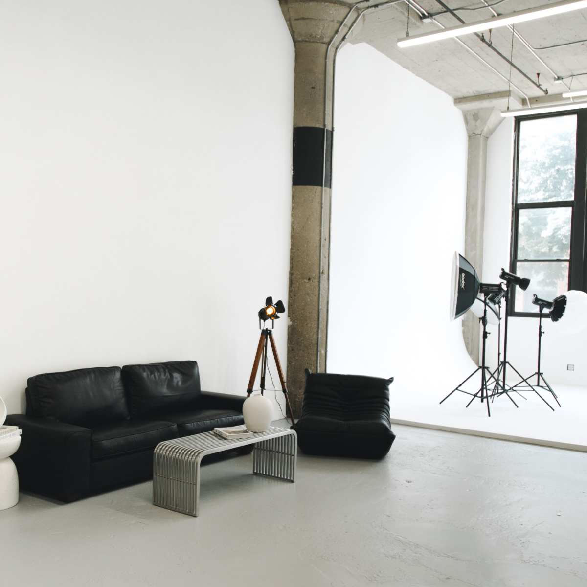 A modern studio space features a black leather sofa, minimalist furniture, and photography equipment with large lights against a bright wall.