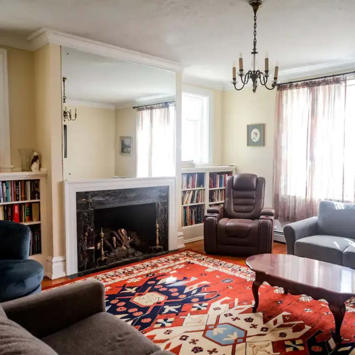 The room features a cozy seating arrangement with a sofa, armchair, and coffee table, arranged around a fireplace, with bookshelves and large windows in the background.