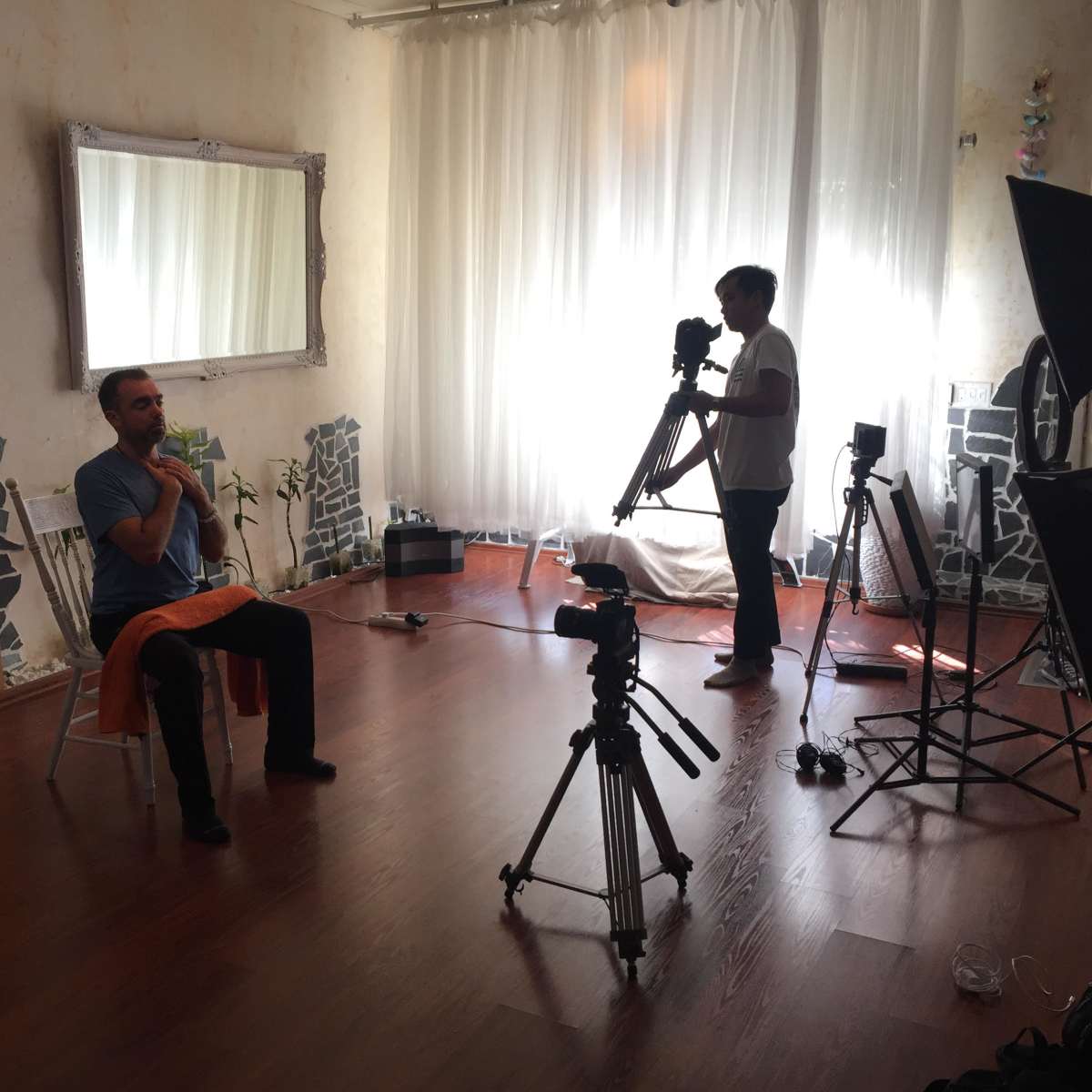 A person sits on a chair in a well-lit room while another person adjusts a camera on a tripod.
