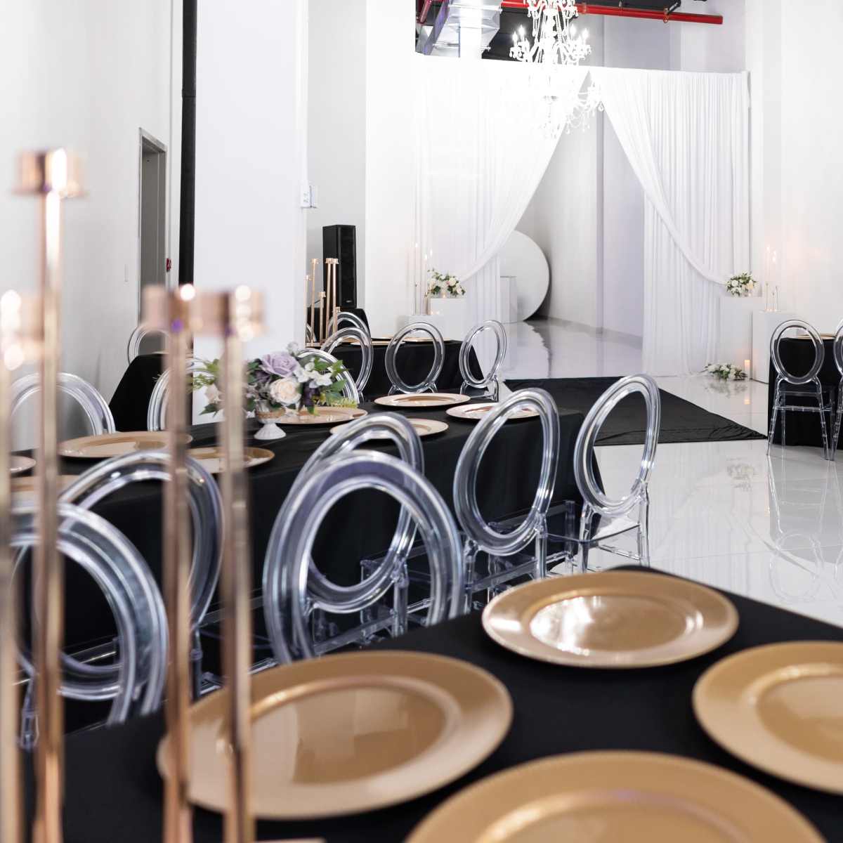 A spacious event venue features black tables set with gold plates and clear acrylic chairs, with white drapes creating a backdrop at one end.