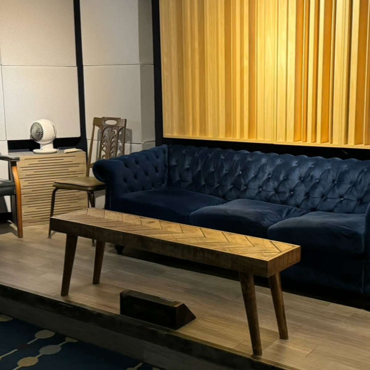 A modern interior with a blue velvet sofa, a wooden coffee table, and two chairs, set against a backdrop of acoustic panels.