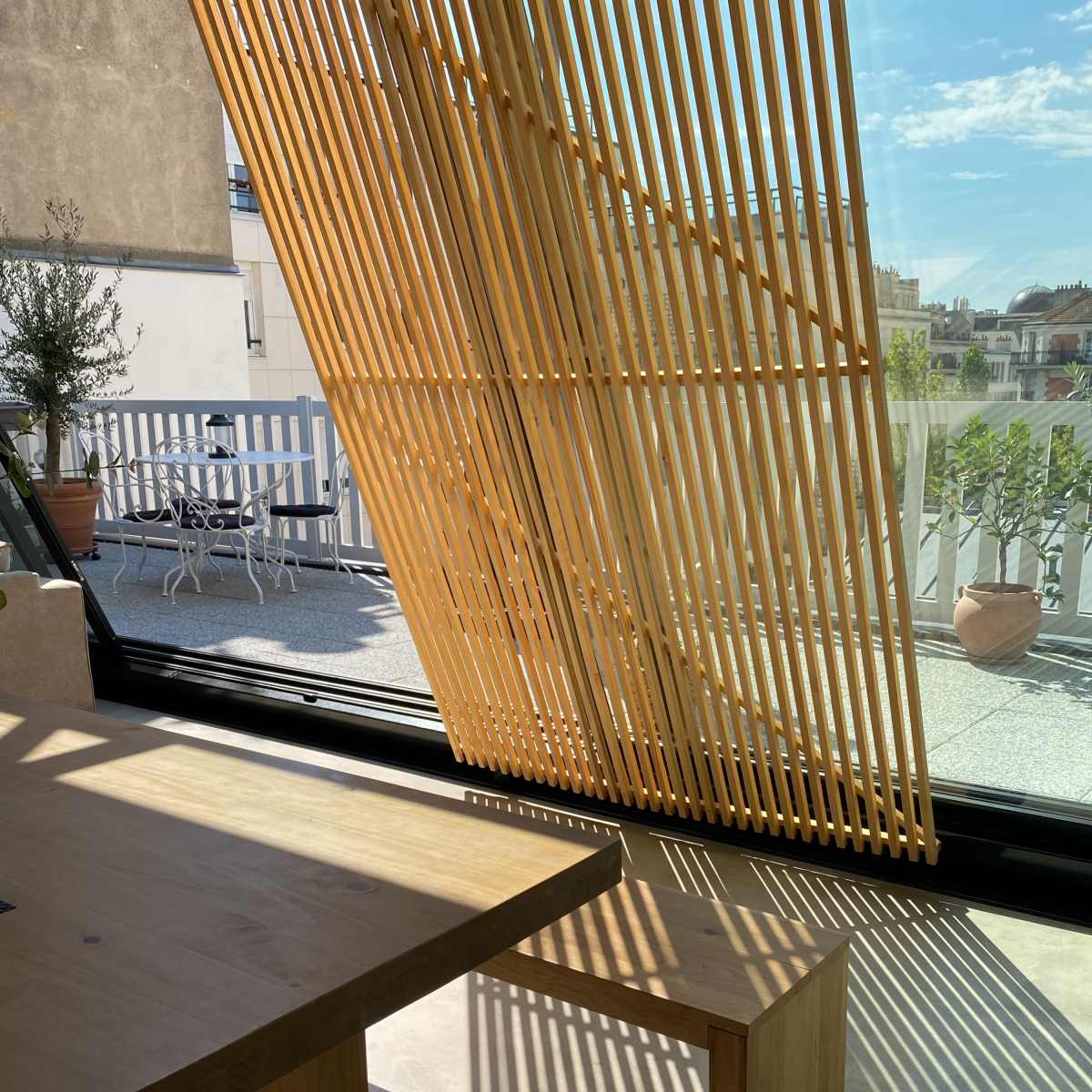 A modern interior space with a wooden table and a bench, illuminated by sunlight filtering through slatted wooden panels, with an outdoor terrace visible in the background.