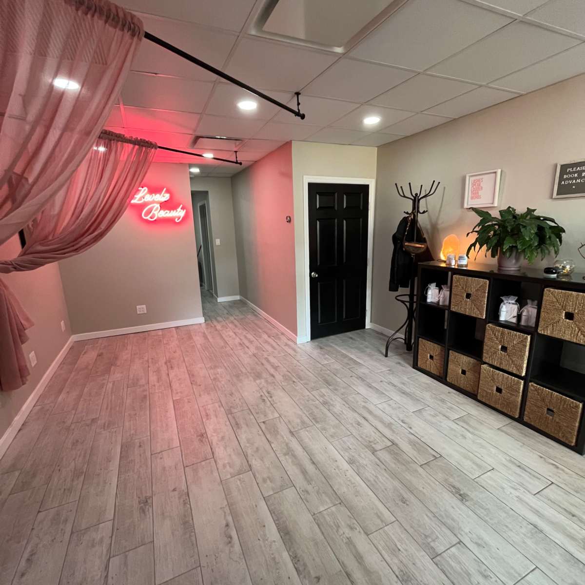 A spacious room with light-colored wooden flooring, a black door, decorative storage boxes, a plant, and a neon sign that reads "Luxe Beauty."