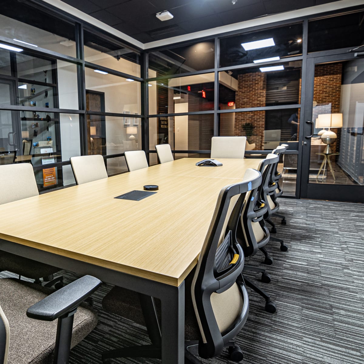 A modern conference room with a long wooden table surrounded by office chairs and large windows.