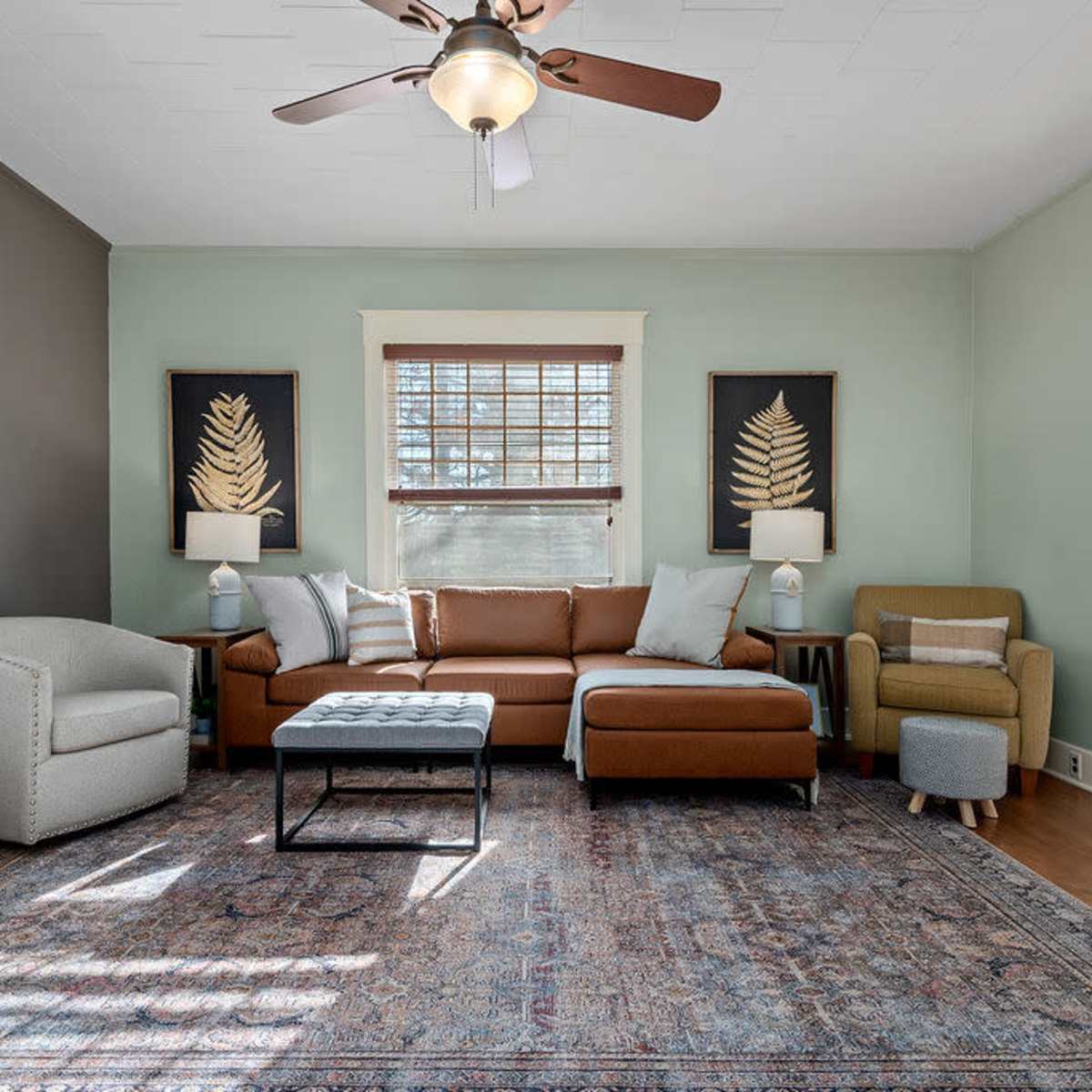 A living room featuring a brown sectional sofa, a gray armchair, and a patterned area rug, with wall art and large windows providing natural light.