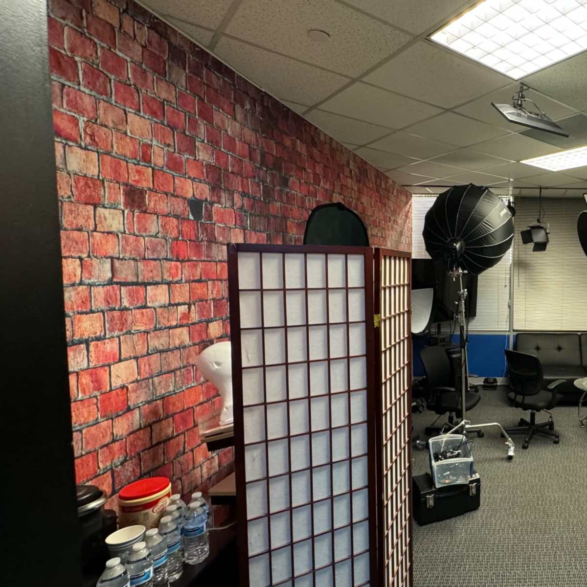A studio setup with a brick-patterned wall, lighting equipment, chairs, and a camera on a tripod in a well-lit space.