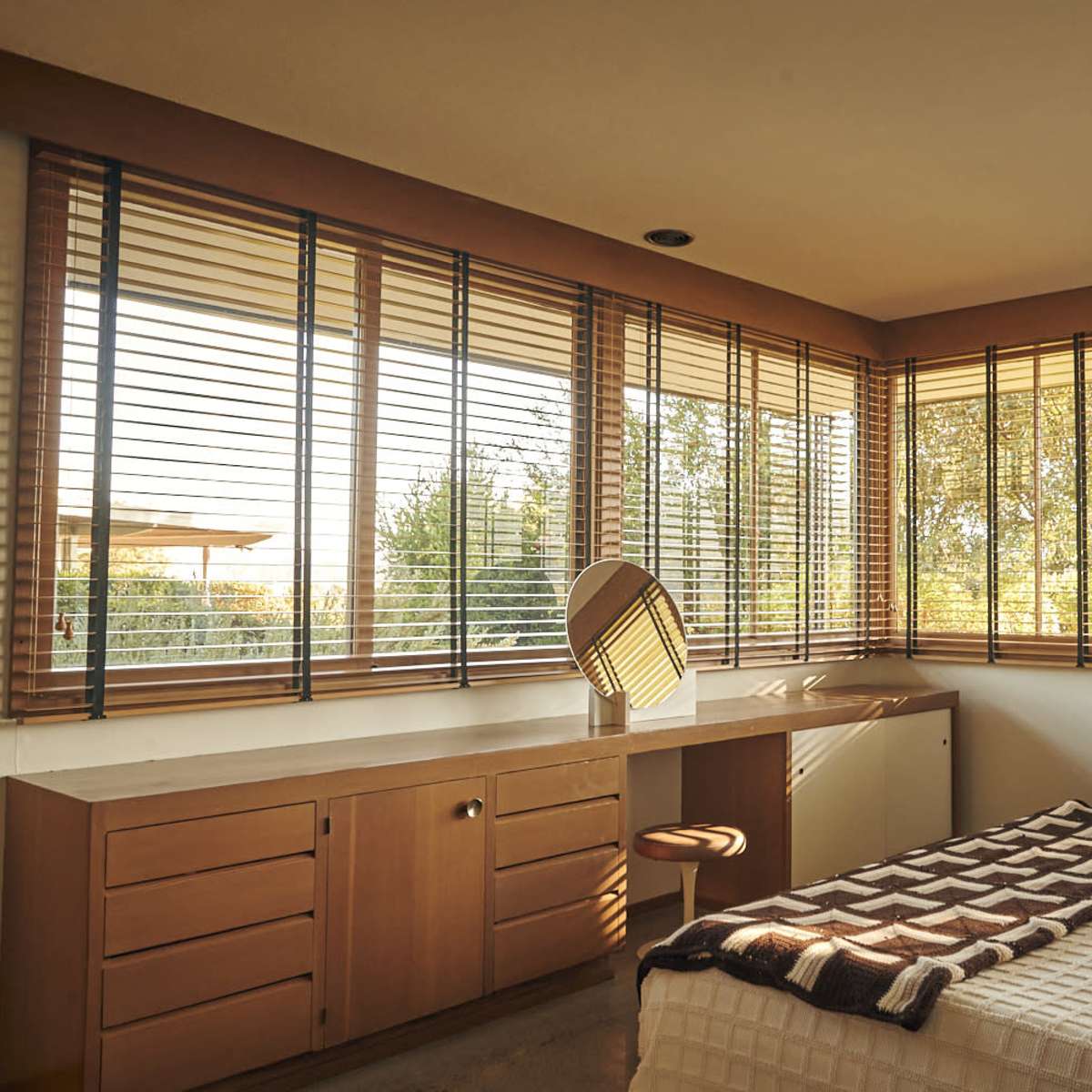 A spacious bedroom with large windows adorned with blinds, a bed with a textured blanket, and a vanity mirror on a wooden desk.
