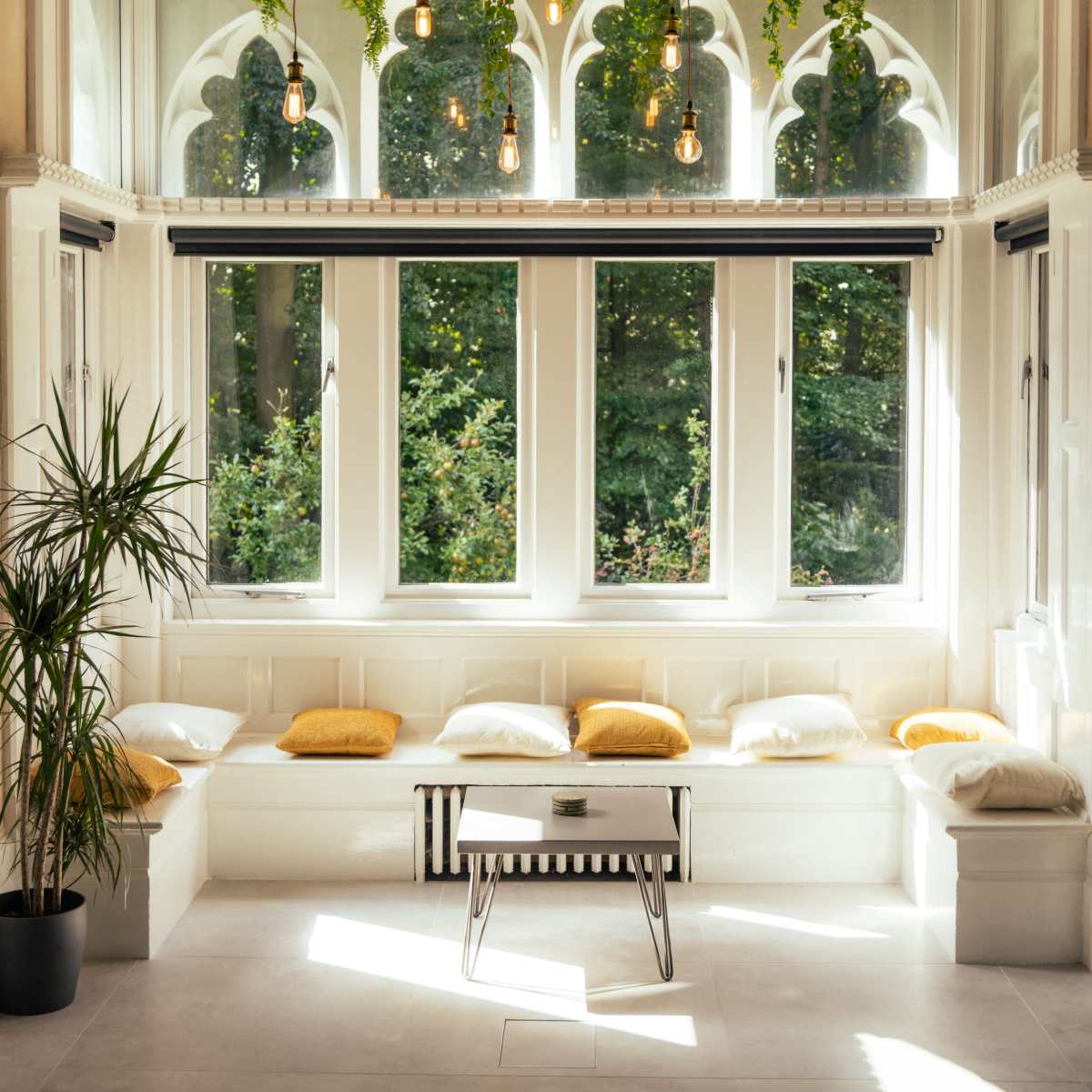 A spacious indoor seating area features large windows, hanging plants, and a central table surrounded by cushions.