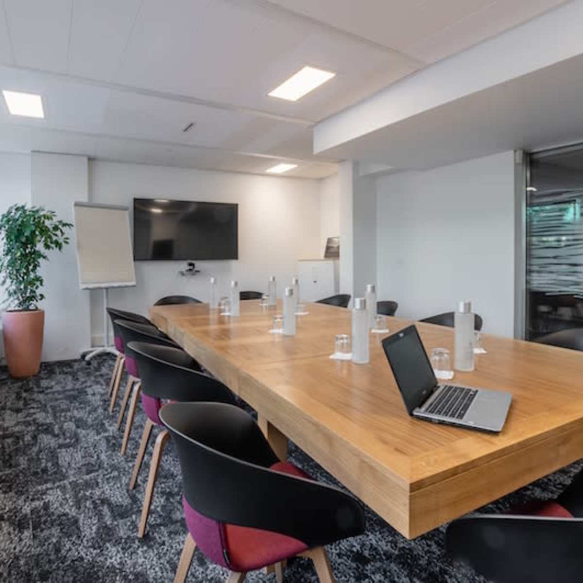 A modern conference room features a large wooden table surrounded by black chairs, with a laptop open on the table and a television screen mounted on the wall.