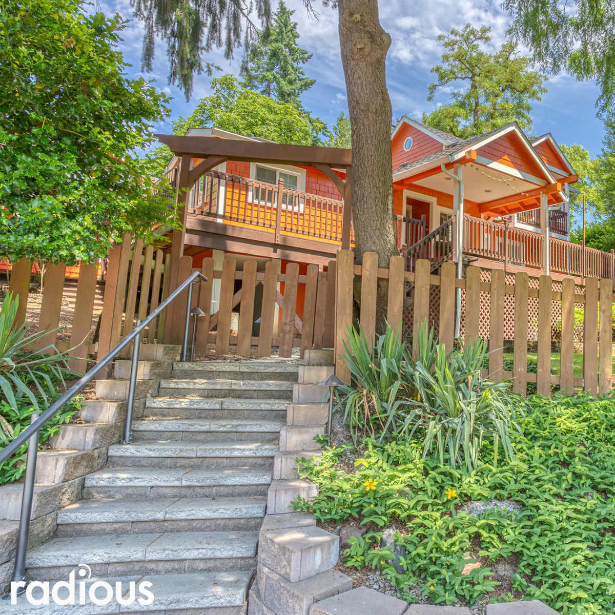 A brightly colored house with a porch and wooden railing is set behind a low fence, surrounded by greenery and trees.