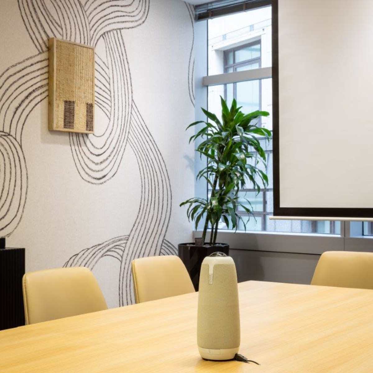 A modern conference room with a round wooden table, a decorative wall with abstract patterns, and a potted plant near a large window.