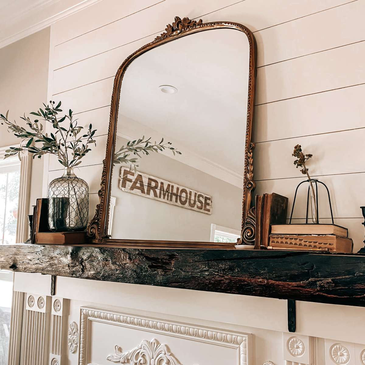 A wooden mantel with a vintage mirror, decorative plants, and various accents is mounted on a white paneled wall.