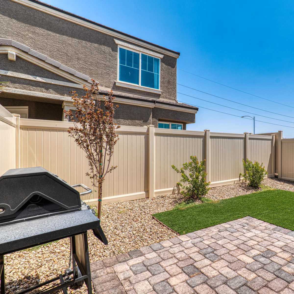 A small backyard with a paved area, artificial grass, and a grill, enclosed by a high fence and adjoining a house.