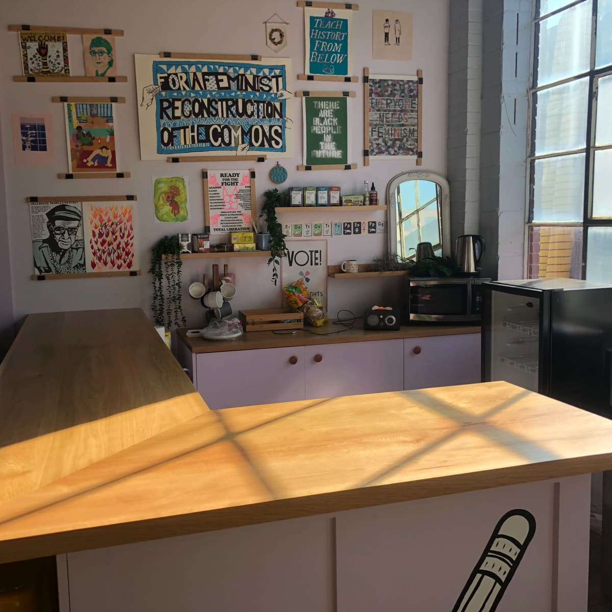 A brightly lit cafe interior with a wooden counter, colorful artwork on the walls, and a small refrigerator.