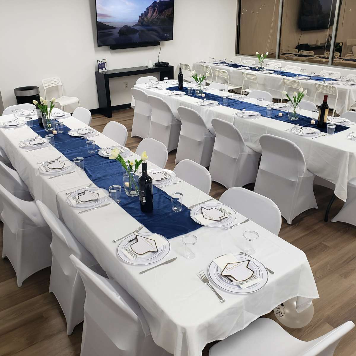 A neatly arranged dining setup in a well-lit room, featuring multiple long tables covered with white tablecloths, plates, and glasses.
