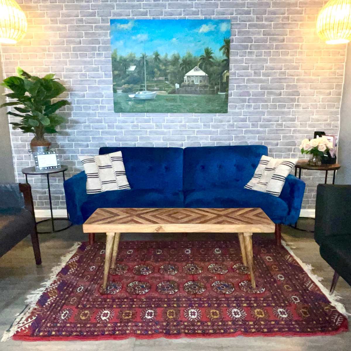 The scene features a living area with a blue couch, two black chairs, a wooden coffee table on a patterned rug, and decor including a painting and potted plants against a brick wall.