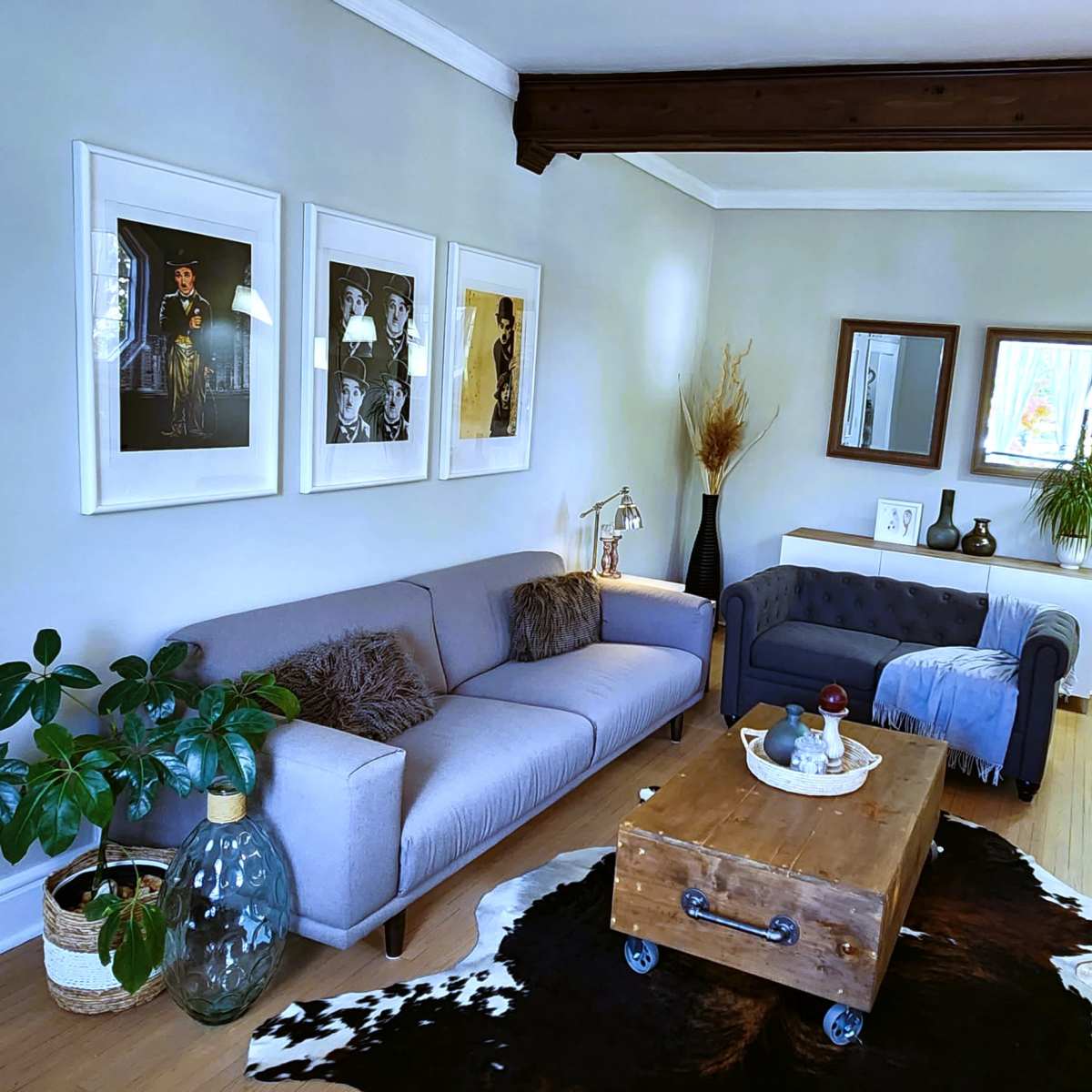 A living room with a gray sofa, a blue tufted armchair, a wooden coffee table, and decorative plants, all set against a light-colored wall with framed artwork.