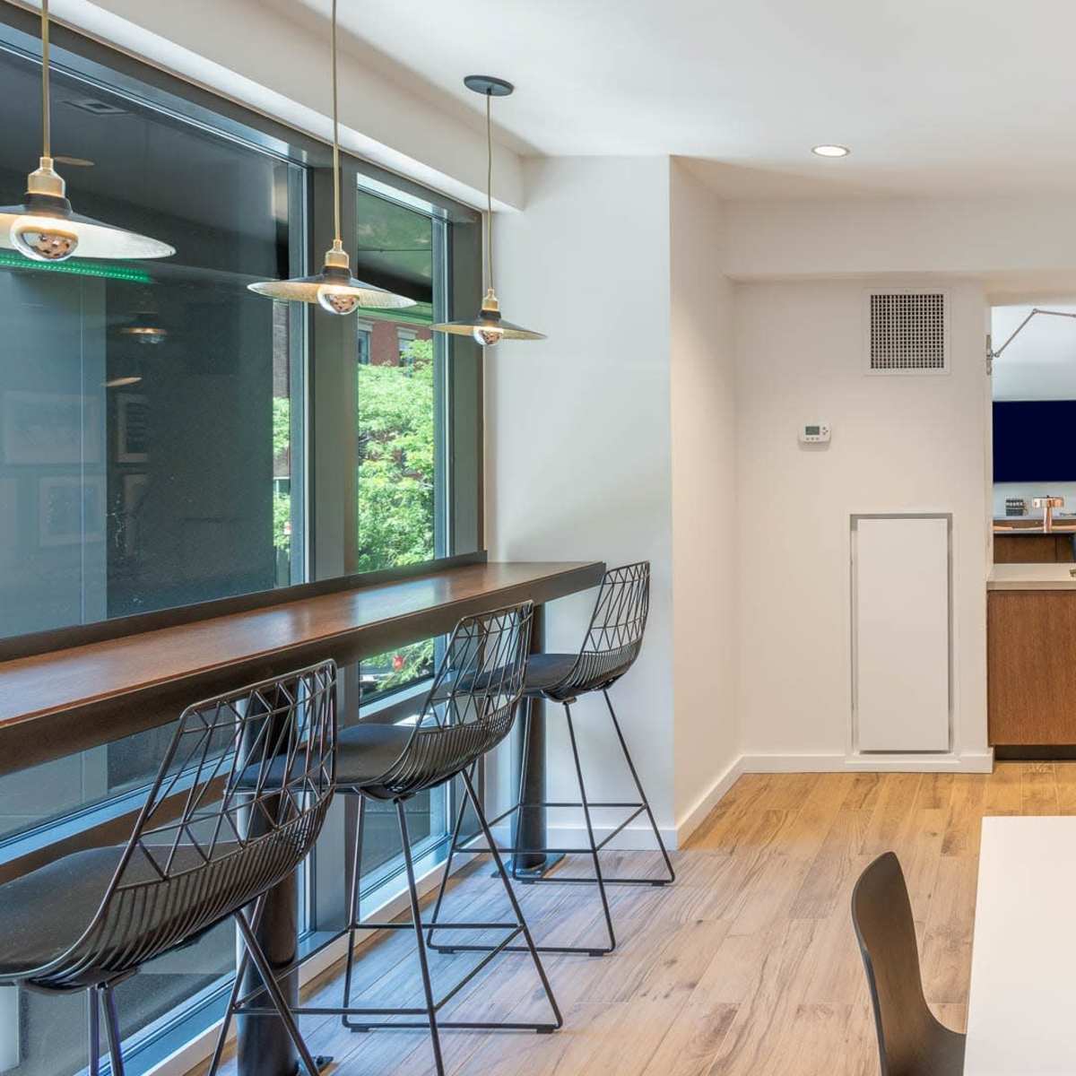 A modern interior space featuring a kitchen area, a dining table, and barstool seating along a window, with wooden flooring and contemporary lighting.