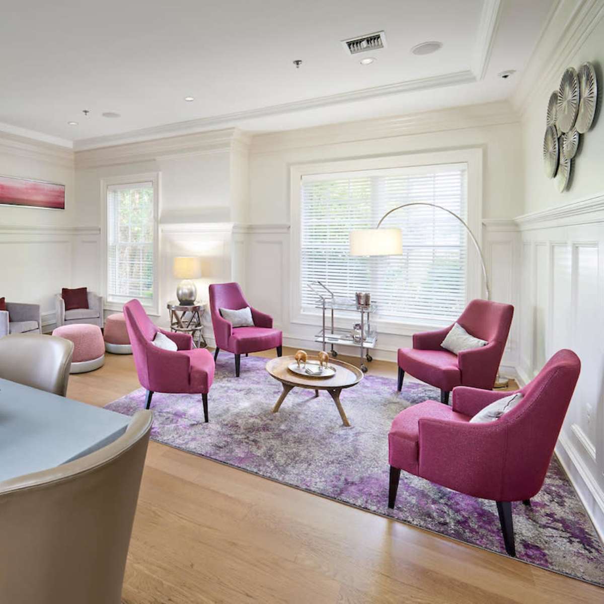 A modern living space features an arrangement of pink armchairs, a round wooden table, a sleek dining table with a bowl of green apples, and large windows providing natural light.