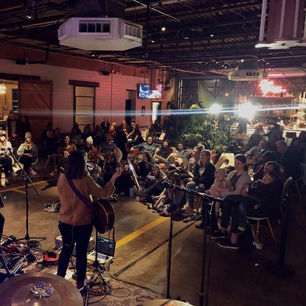 Two musicians perform on stage in a dimly lit venue while an audience sits facing them.