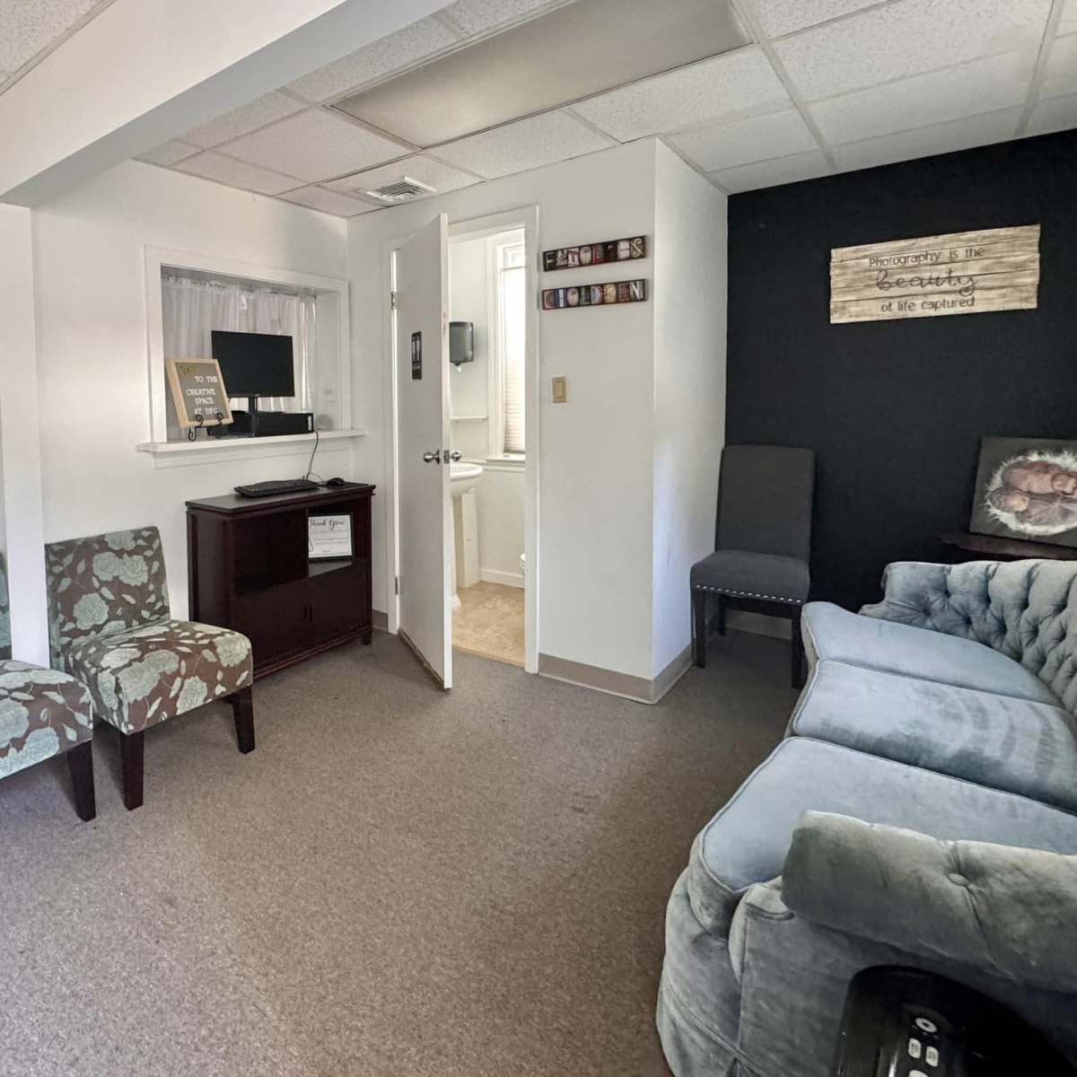 A small living area with two patterned chairs, a wooden cabinet, and a gray sofa, with a doorway leading to a bathroom and a wall displaying decorations.