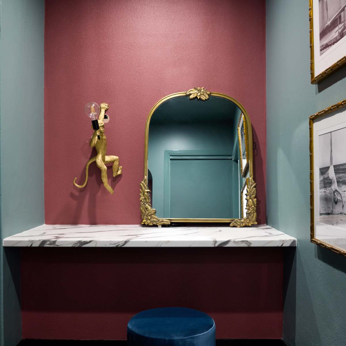 A small bathroom with a maroon wall, a marble-topped vanity, an ornate gold-framed mirror, and a blue velvet stool.