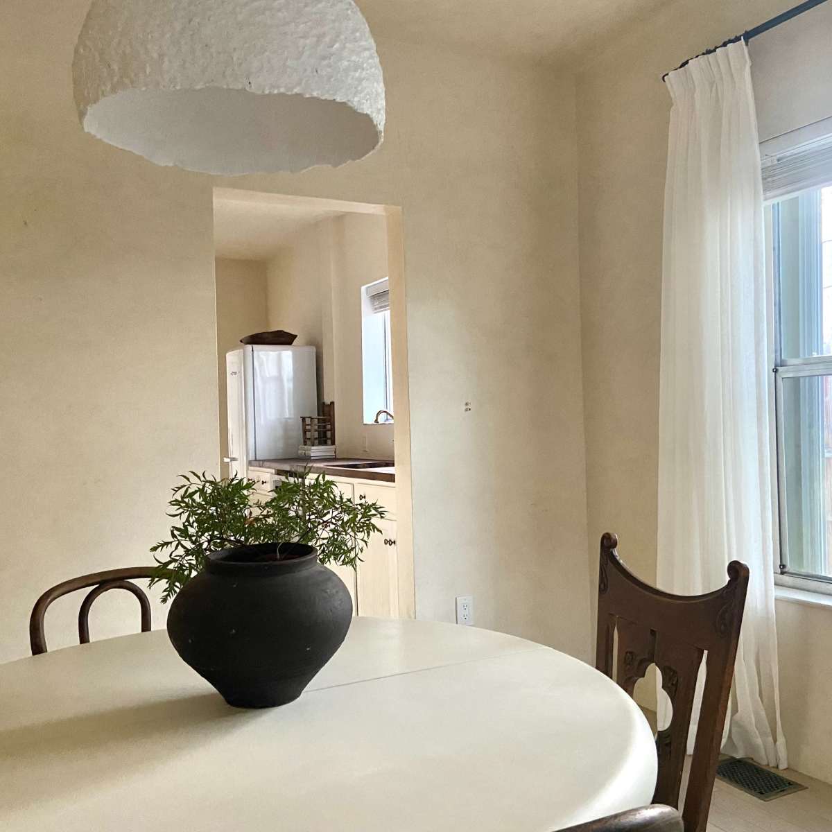 A round dining table with a dark pot of green foliage sits under a pendant lamp in a light-colored room next to a window with sheer curtains.