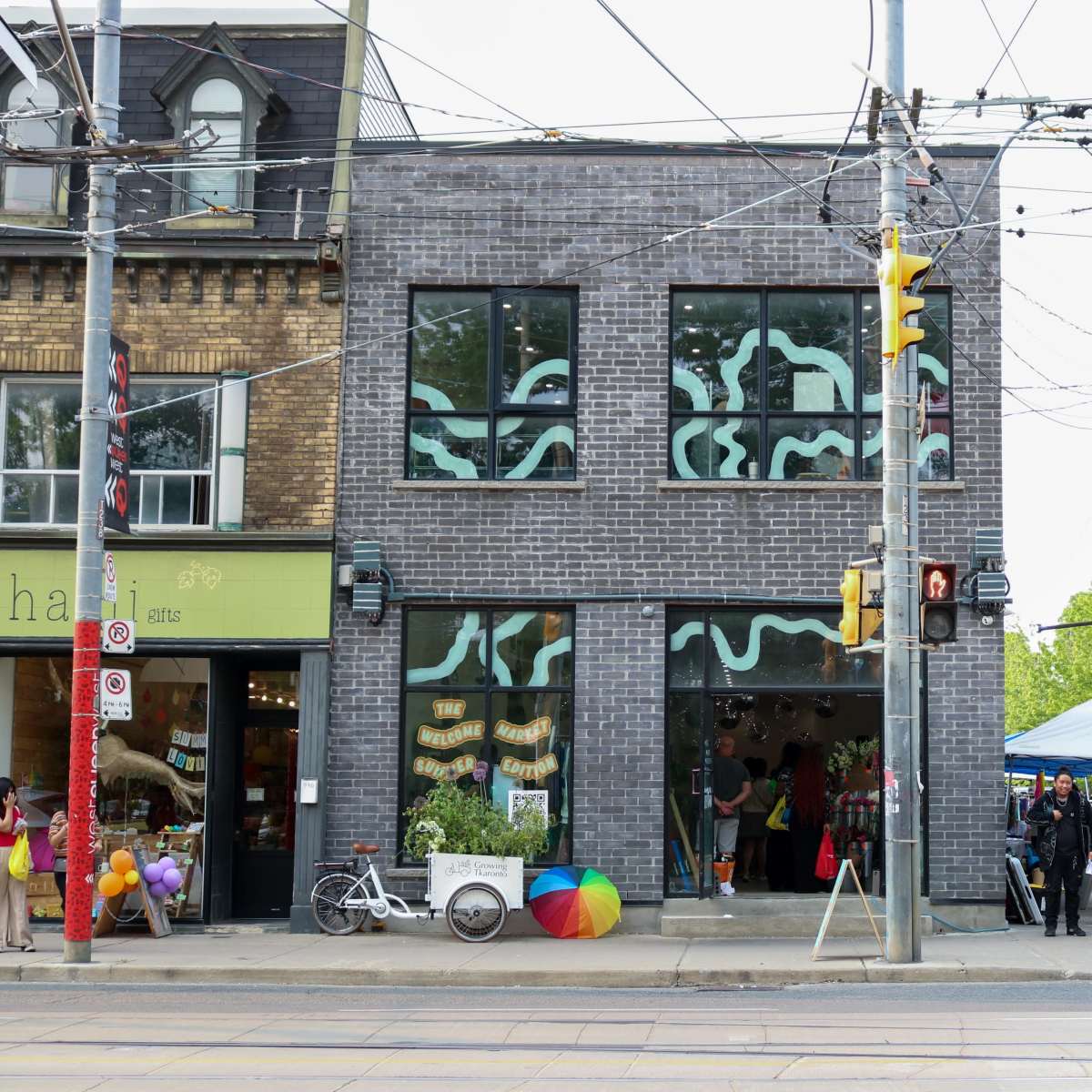 The scene features a modern brick building with large windows displaying colorful decorations, while pedestrians gather outside near a street with cars and a pop-up tent.