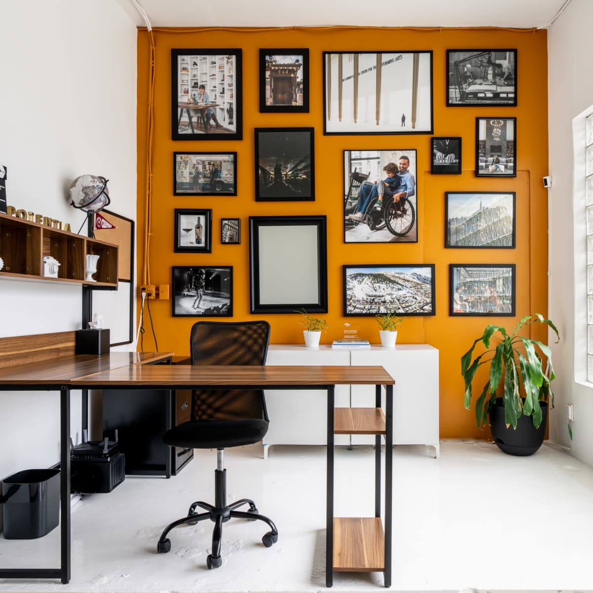 A modern home office features a wooden desk and a black chair facing a gallery wall of framed art against an orange backdrop.