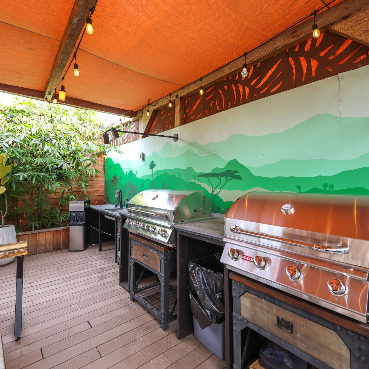 A covered outdoor grilling area with stainless steel grills, wooden countertops, and a mural of green hills on the wall.