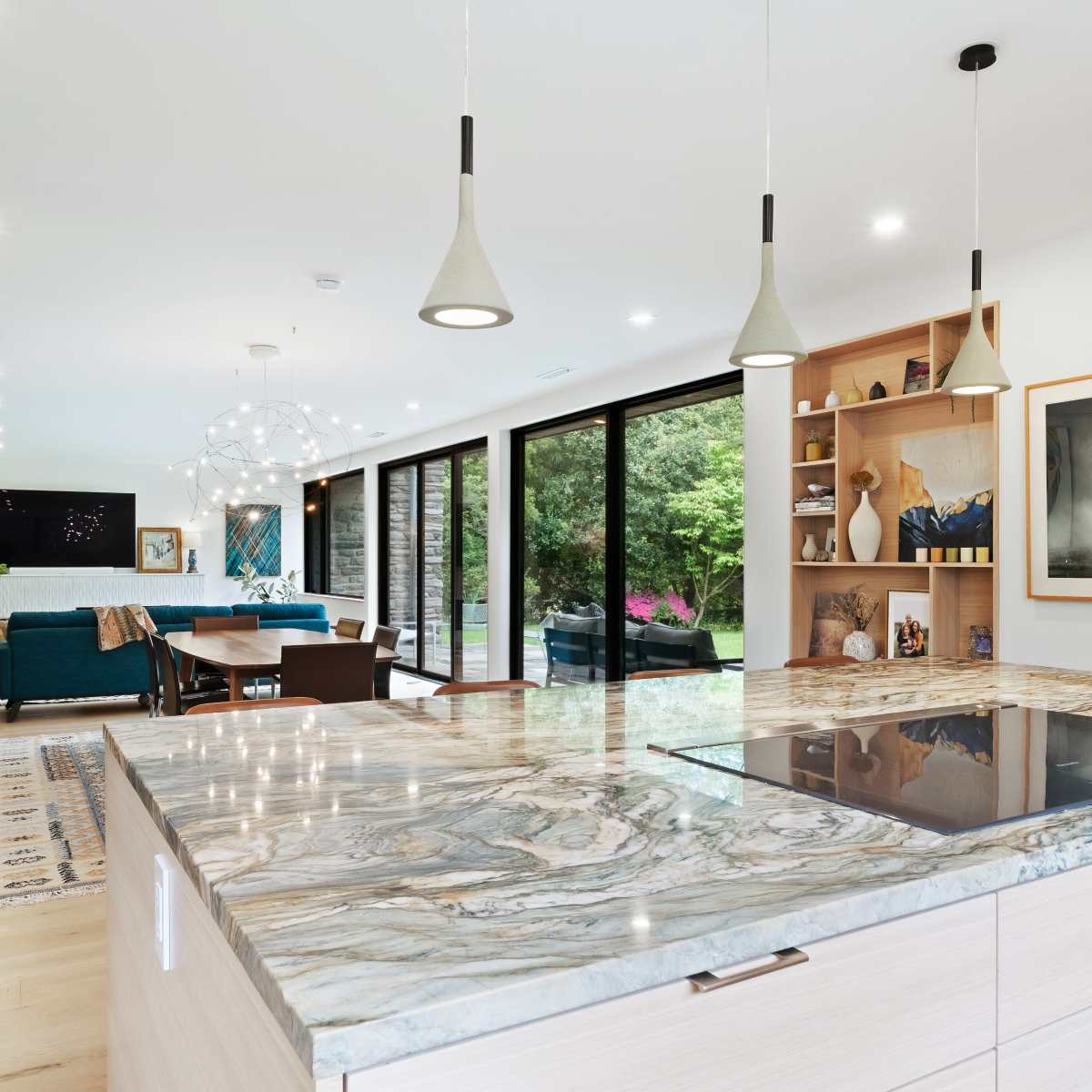 A modern kitchen with a large island featuring a cooktop, overlooking a living area with large windows revealing a view of greenery outside.