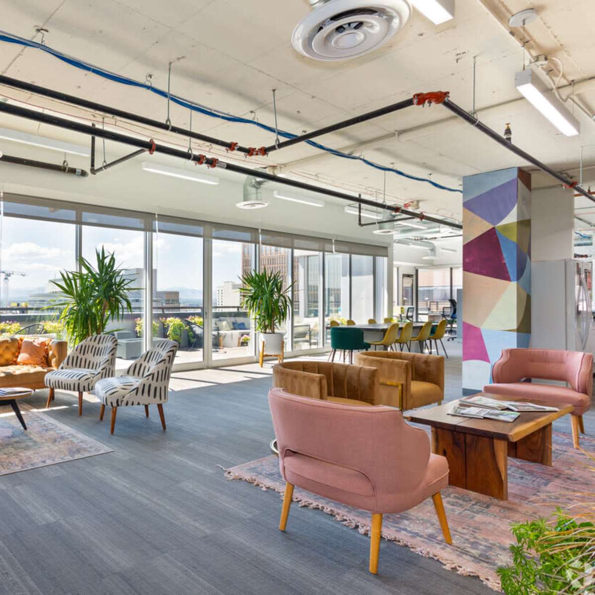 A modern office lounge area with various seating arrangements, large windows, and plants, creating a bright and open atmosphere.