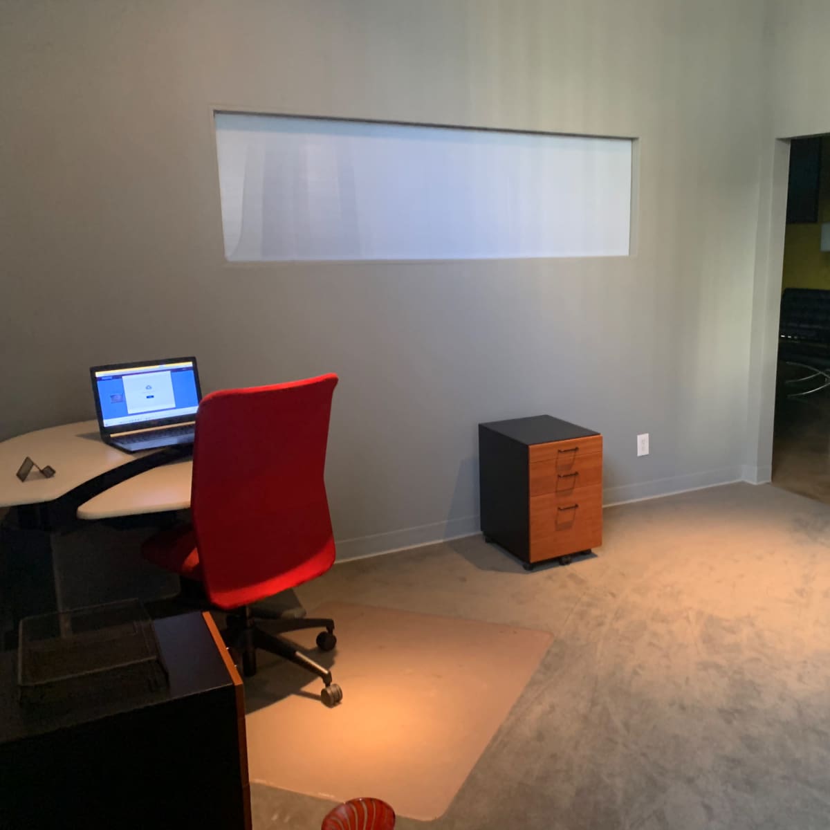 A sparse office space with a desk, a laptop, a red chair, a filing cabinet, and a wooden chair in a dimly lit corner.
