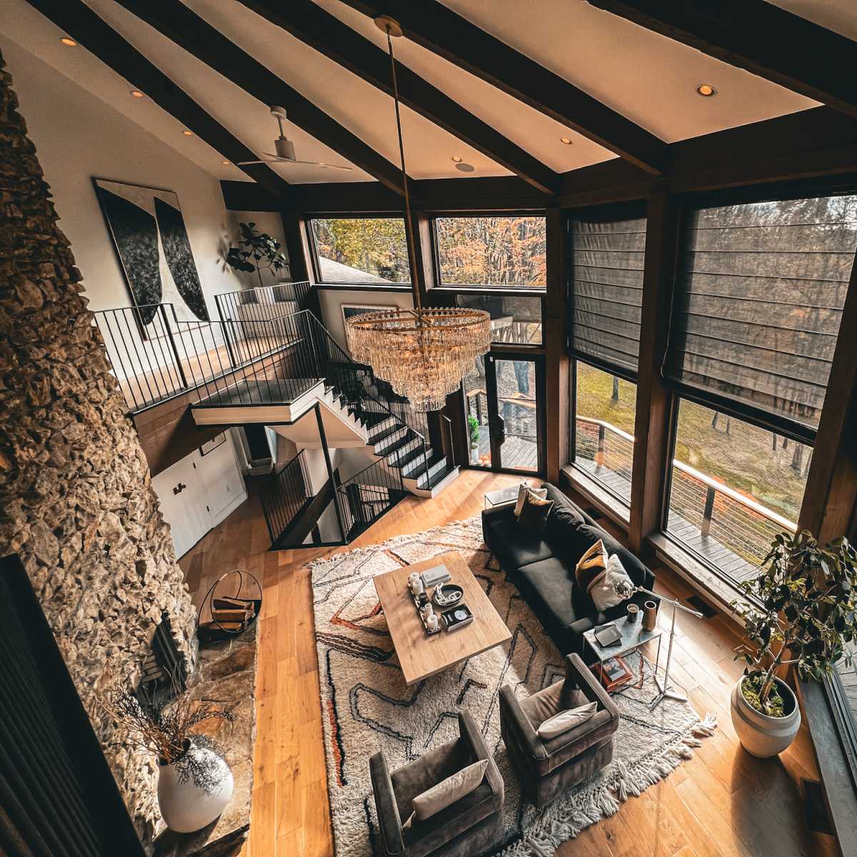 A spacious, modern living room with large windows, a stone fireplace, and a staircase leading to an upper level.
