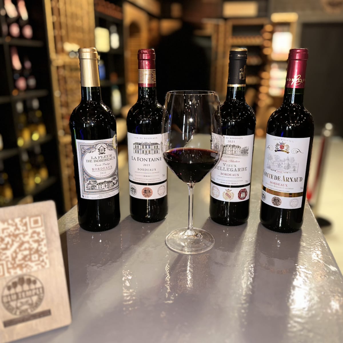 Four wine bottles arranged next to a filled wine glass on a countertop, with a QR code sign displayed in front.