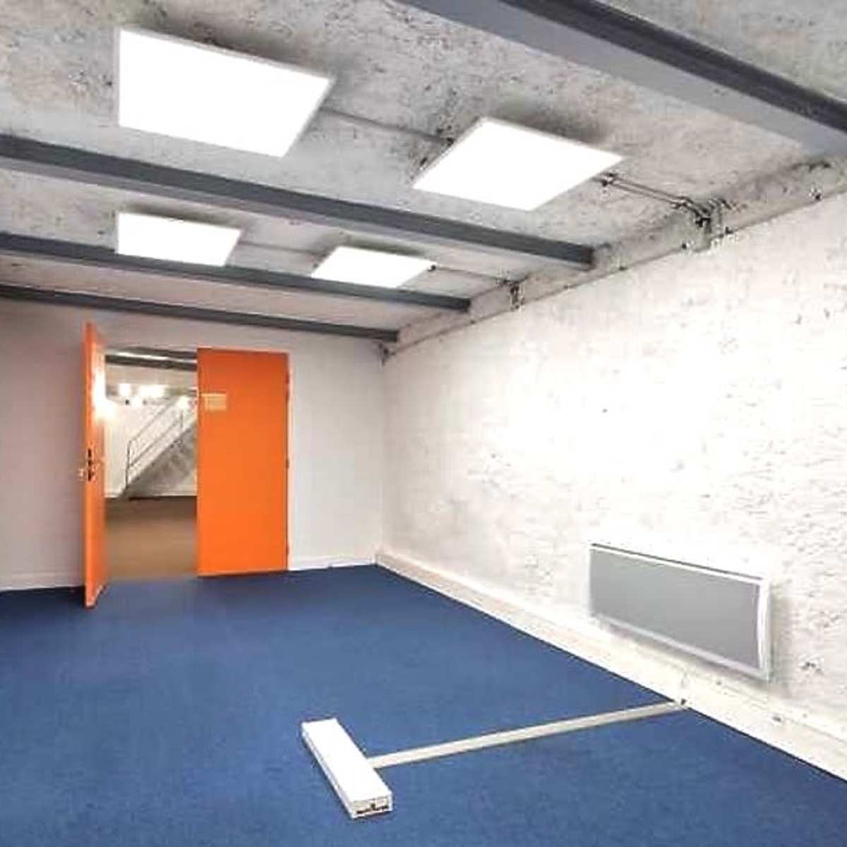 A sparsely furnished room with a blue carpet, white walls, and an orange door leading to another space.