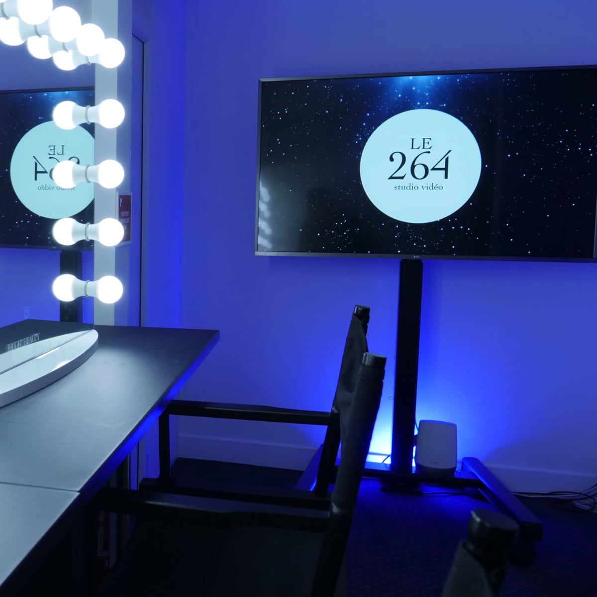 A well-lit makeup vanity with large circular lights and a nearby screen displaying the text "LE 264 studio vidéo."