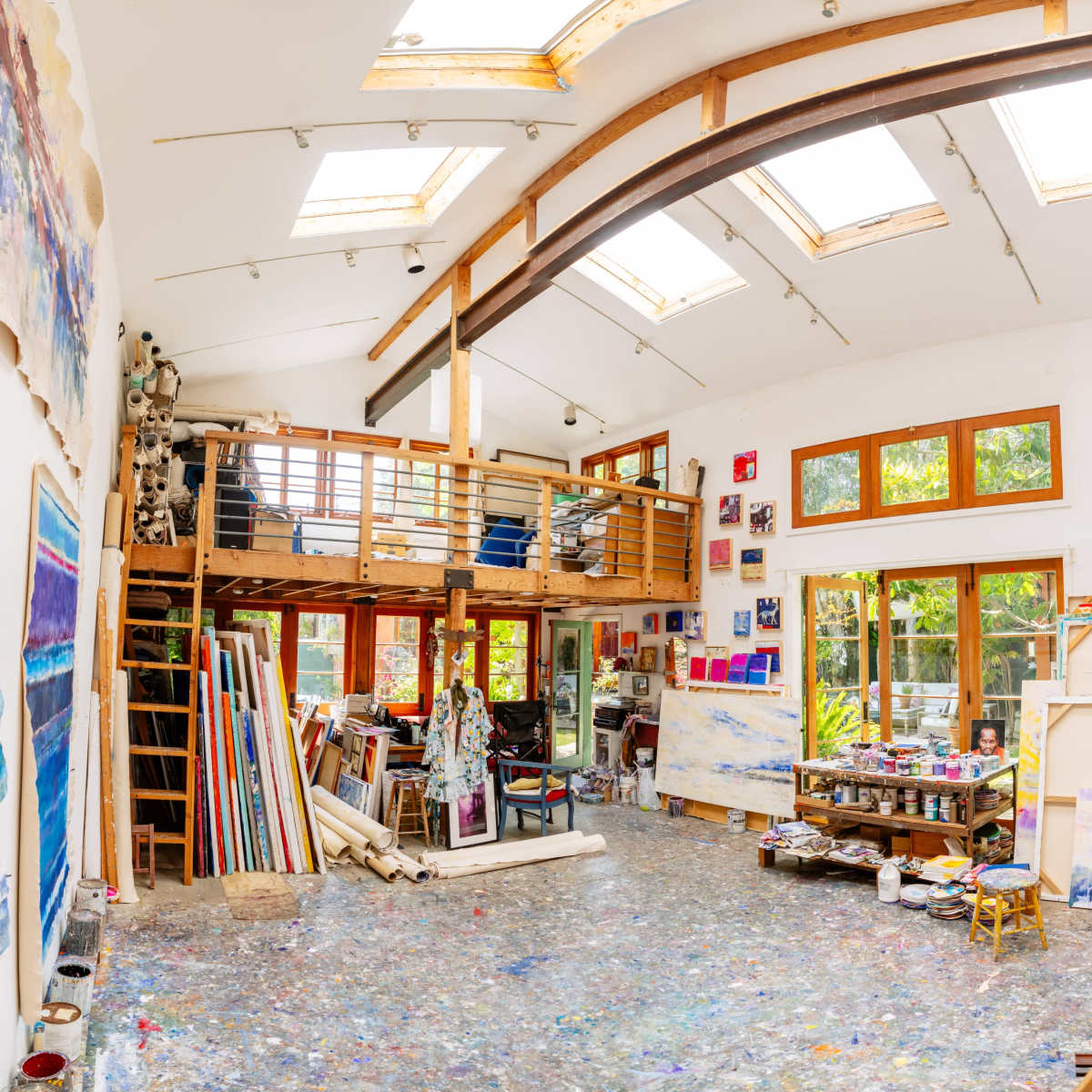 An artist's studio with a high ceiling, large windows, and an open space filled with canvases, paint supplies, and a loft area.