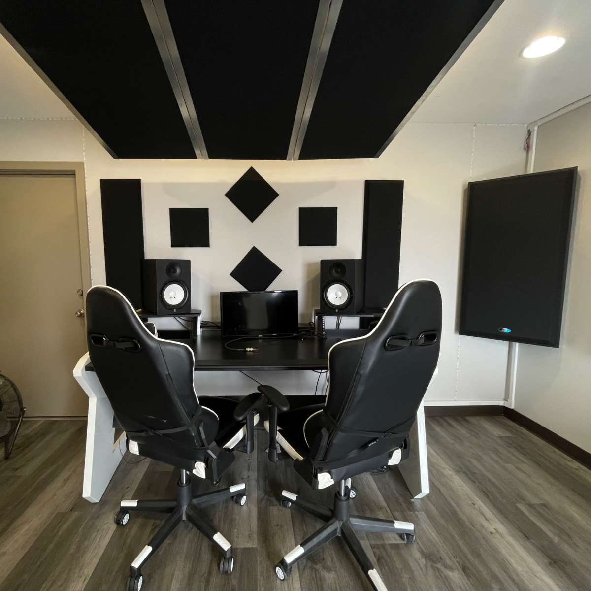 A modern recording studio with two black office chairs facing a desk equipped with speakers and a computer monitor, surrounded by sound-absorbing panels.