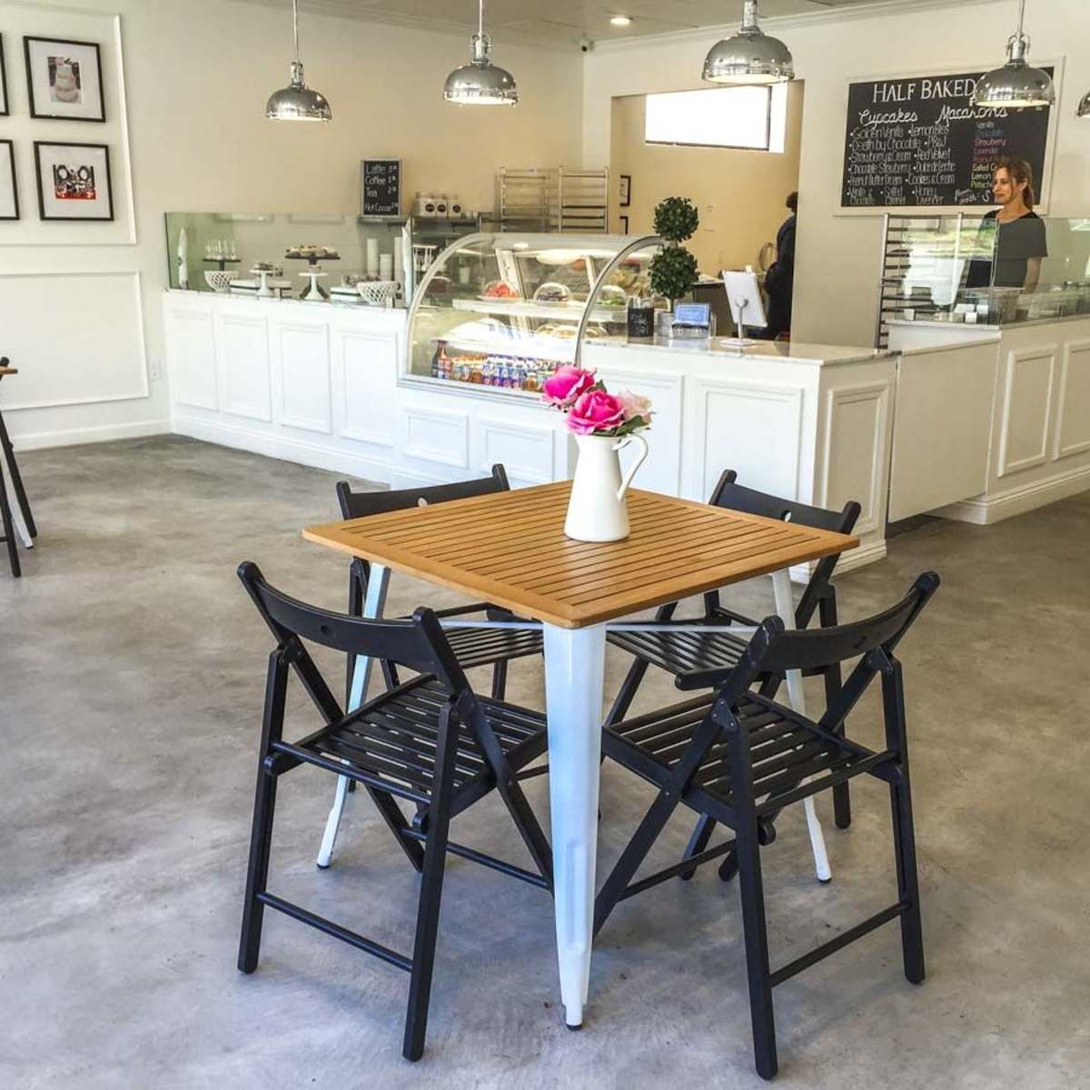 A bright and modern cafe with a wooden table and black folding chairs arranged in the center, complemented by a glass display case of desserts in the background.