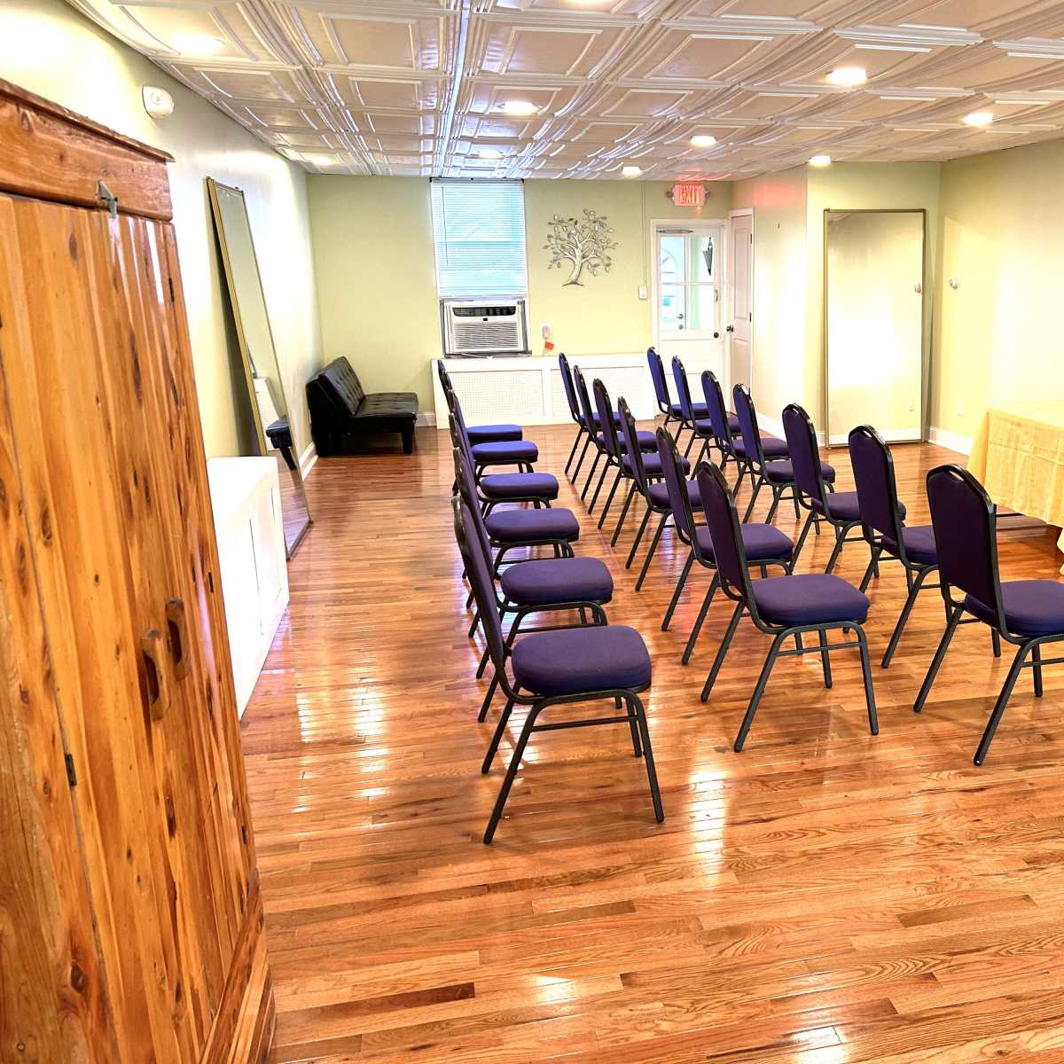 A well-lit room with polished wooden floors, featuring several rows of chairs facing a table in the front.