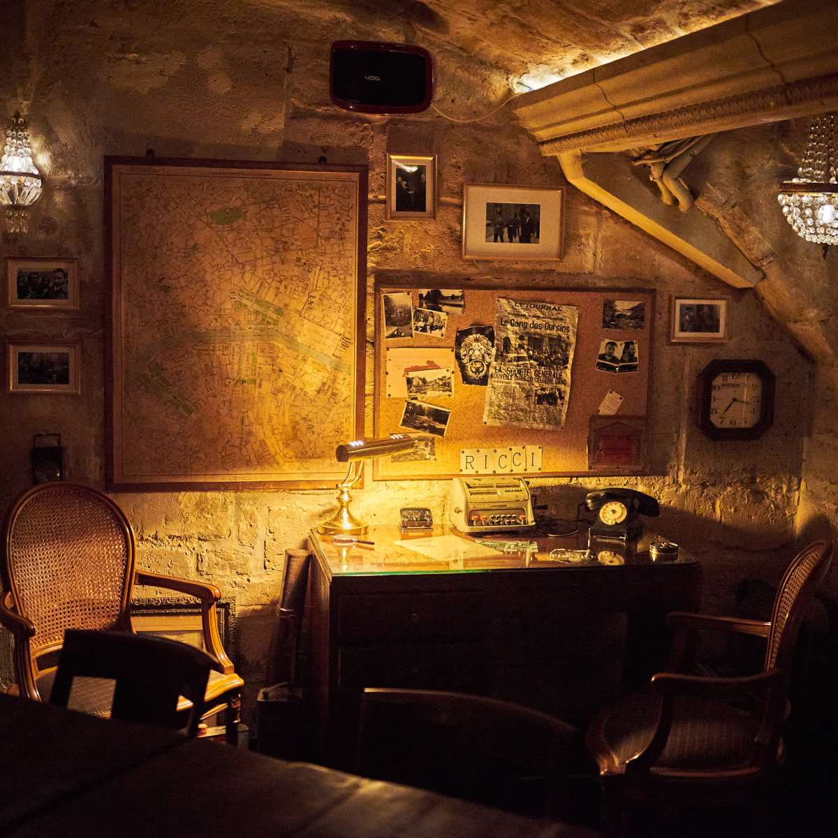 A dimly lit room features a wooden desk with a vintage lamp, old photographs, a rotary phone, and two wicker chairs against a stone wall adorned with a large map and framed images.