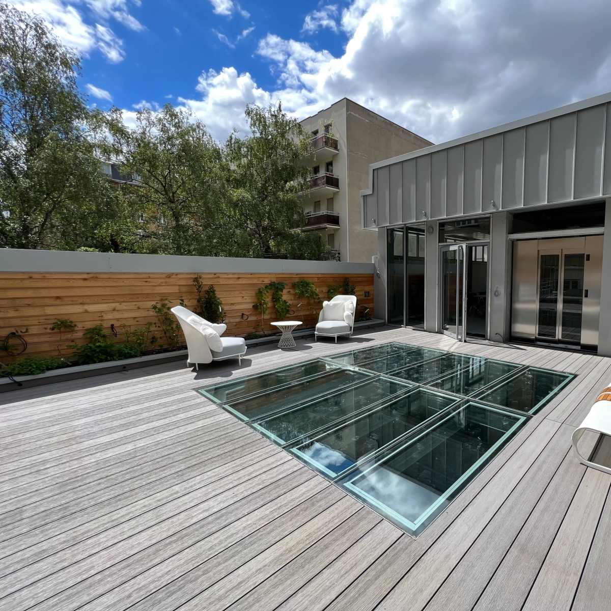 A modern rooftop terrace features a glass floor section, two white chairs, a small table, and surrounding greenery.