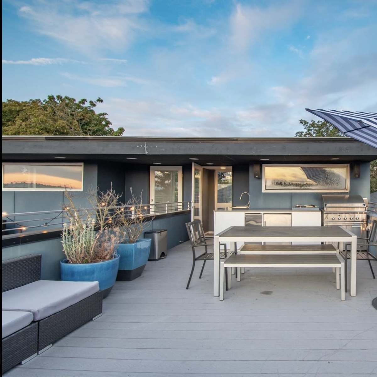 A rooftop terrace features a dining table, an outdoor kitchen, potted plants, and a striped umbrella against a blue sky.