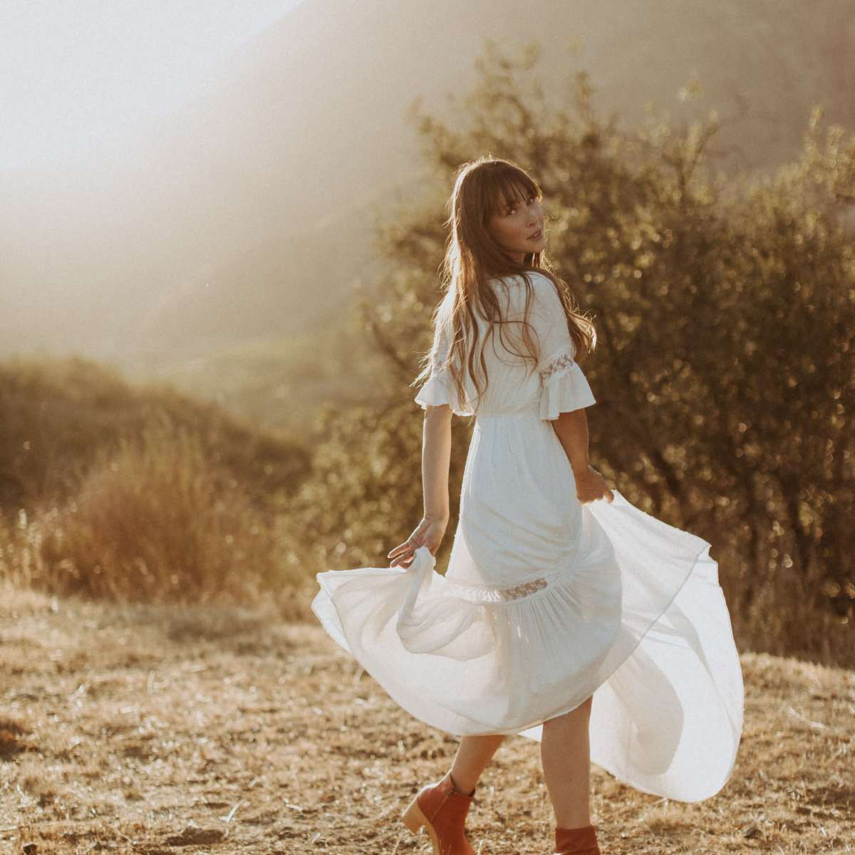 A woman in a white dress twirls in a sunlit field with mountains and shrubs in the background.