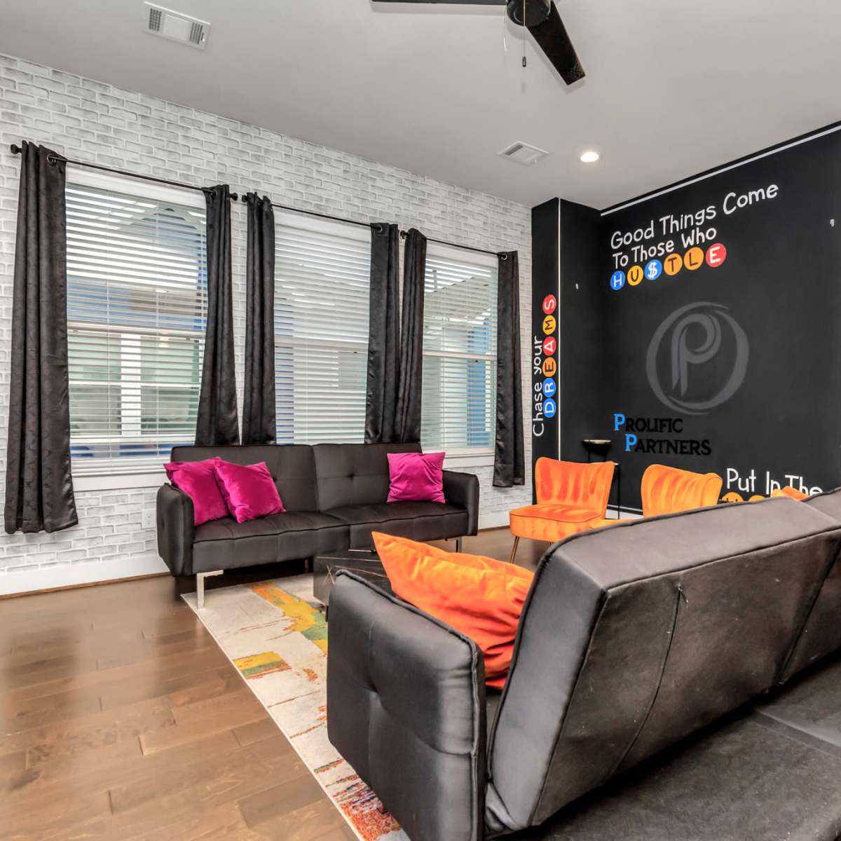 A modern living room with a black sofa, colorful chairs, a patterned rug, and a feature wall with motivational text and a chalkboard area.