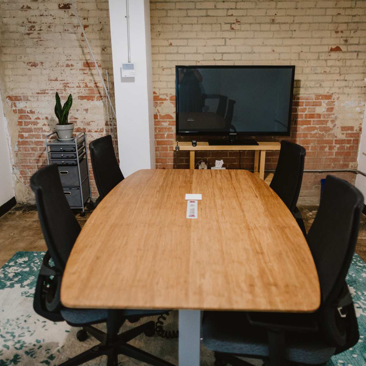 A conference room features a large wooden table surrounded by ergonomic chairs, with a television mounted on a wall and a potted plant nearby.
