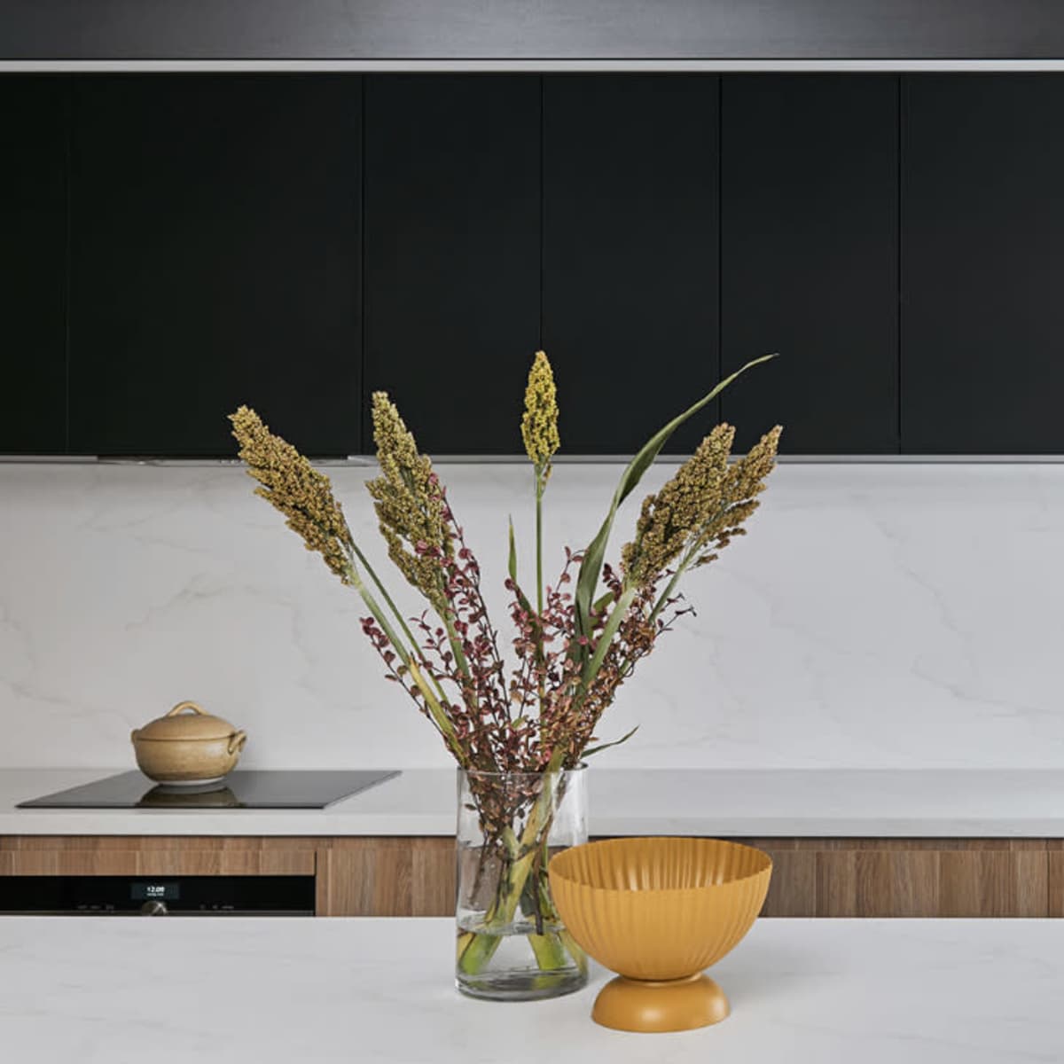 A modern kitchen features a marble countertop with a clear vase holding dried flowers and a yellow bowl, set against black cabinets and a minimalist sink.