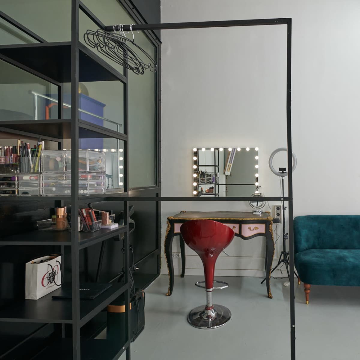 A studio space featuring a black metal clothing rack, a teal sofa, a vanity mirror with lights, and a red stool beside a makeup storage unit.